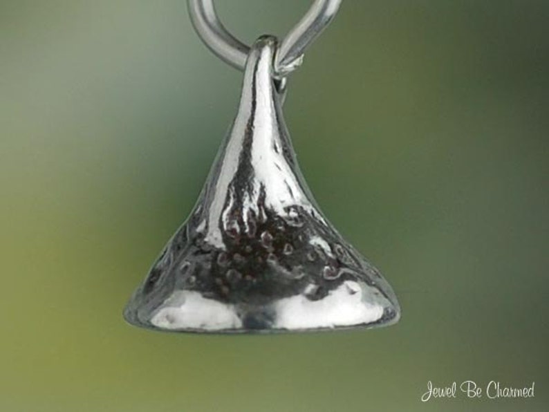May include: A silver-colored pendant in the shape of a Hershey's Kiss, with a textured surface. The pendant is suspended from a silver loop. The background is a soft gradient of green and yellow.