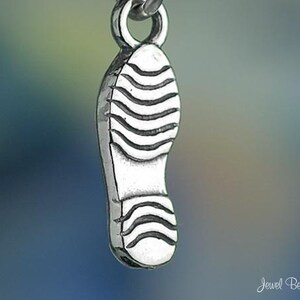 May include: A silver charm in the shape of a shoe with a textured sole. The charm is designed to be added to a bracelet or necklace.
