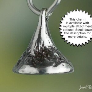May include: A silver-colored charm in the shape of a Hershey's Kiss, suspended from a silver loop. The charm has a textured surface and is set against a blurred green background. Text on the image states, "This charm is available with multiple attachment options! Scroll down the description for more details."