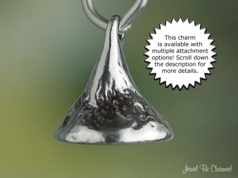 May include: A silver-colored charm in the shape of a Hershey's Kiss, suspended from a silver loop. The charm has a textured surface and is set against a blurred green background. Text on the image states, "This charm is available with multiple attachment options! Scroll down the description for more details."