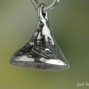 May include: A silver-colored pendant in the shape of a Hershey's Kiss. The pendant has a textured surface and is suspended from a small loop. The background is a soft green and yellow gradient.
