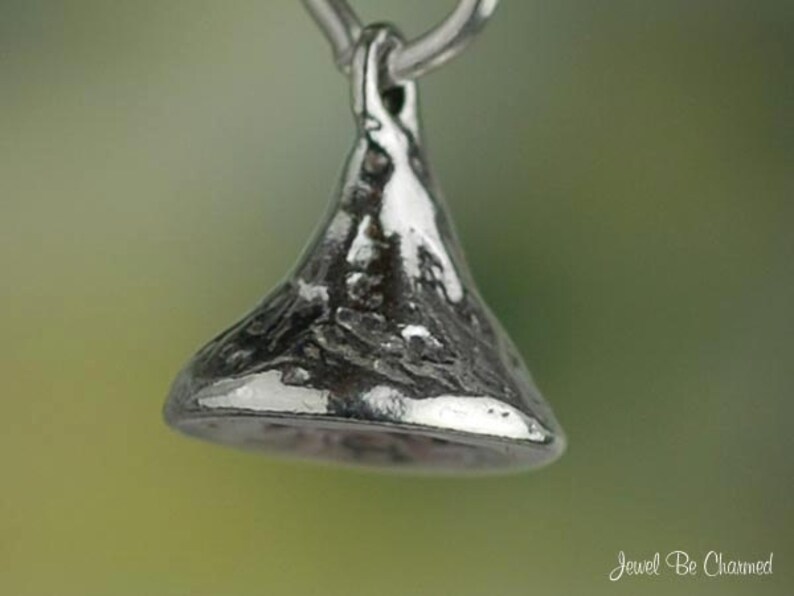 May include: A silver-colored pendant in the shape of a Hershey's Kiss. The pendant has a textured surface and is suspended from a small loop. The background is a soft green and yellow gradient.