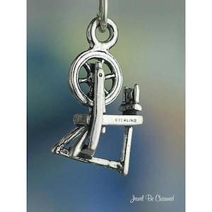 May include: A silver-colored charm in the shape of a spinning wheel. The charm has a wheel, a frame, and other details. The word "STERLING" is stamped on the frame. The charm is suspended from a small loop.