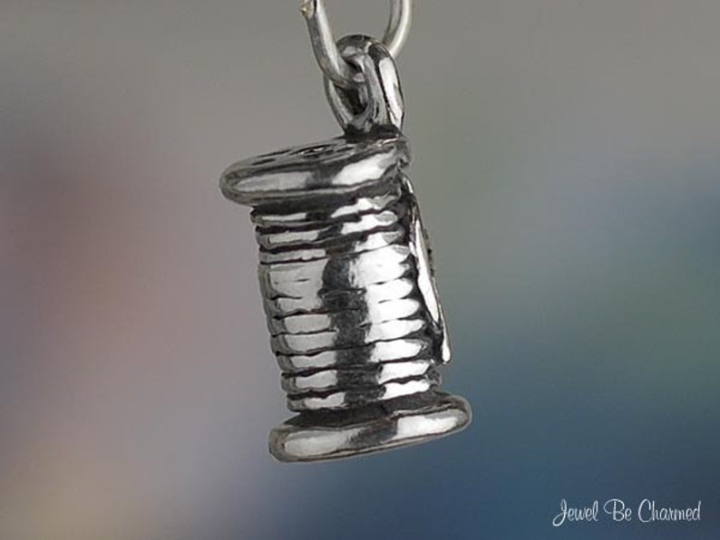 Puede incluir: Un charm de plata con forma de carrete de hilo. El charm est&aacute; detallado con una superficie texturizada y un bucle para sujetarlo a una pulsera o collar.