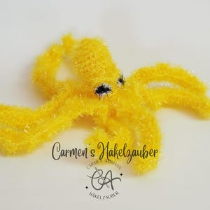 May include: A handmade, bright yellow crocheted octopus with black eyes. The octopus has eight textured tentacles and is displayed on a white background. The text "Carmen's Hakelzauber" is visible.