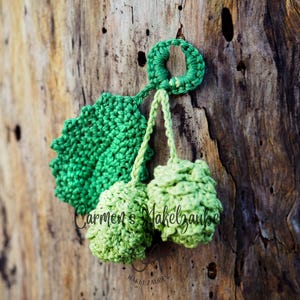 Pode incluir: Um ornamento de crochê verde feito à mão, semelhante a um raminho de cerejas, com uma folha e duas cerejas redondas. O ornamento está pendurado em um fundo de madeira marrom texturizada. O texto "Carmen's Makelzauber" é visível.