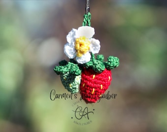 May include: Crocheted strawberry and flower ornament. The red strawberry has yellow accents, green leaves, and a white and yellow flower. The ornament is suspended against a blurred green background. The text "Carmen's Hakelzauber" is visible.