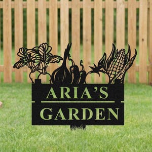 May include: Black metal garden sign with the text "ARIA'S GARDEN" and a decorative top featuring silhouettes of vegetables, including garlic, peppers, and corn. The sign is placed in a grassy yard with a wooden fence in the background.