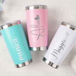 以下が含まれることがあります： ターコイズ、ピンク、白の3色の、パーソナライズされたステンレススチールタンブラー。各タンブラーは銀色の蓋と底があり、筆記体で名前が書かれています。ピンクのタンブラーにはハートのグラフィックがあります。ターコイズのタンブラーには「Monica」、ピンクには「Alice」、白には「Maggie」と書かれています。