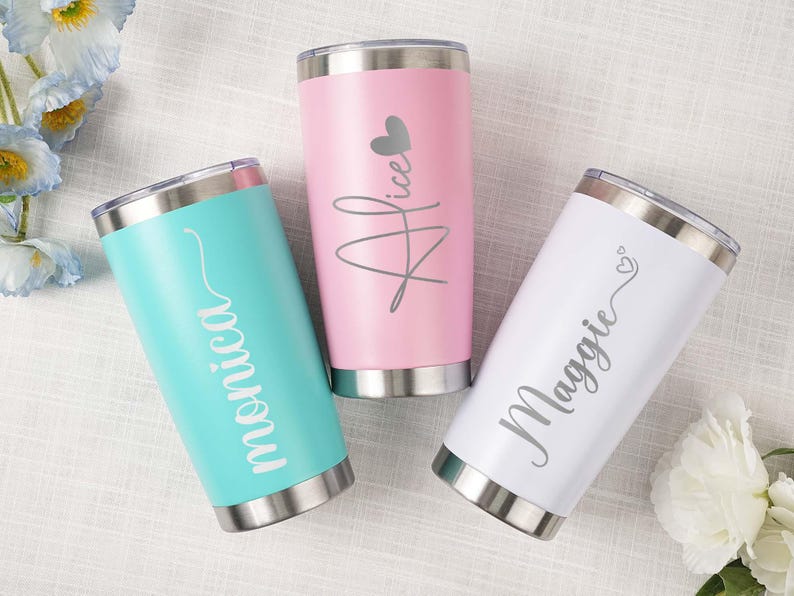 May include: Three personalized stainless steel tumblers in teal, pink, and white. Each tumbler has a silver lid and base, with a name in a cursive font. The pink tumbler has a heart graphic. The teal tumbler says "Monica", the pink one says "Alice", and the white one says "Maggie".
