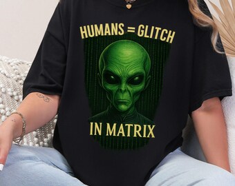 May include: Black t-shirt featuring a green alien face with the text "HUMANS = GLITCH IN MATRIX" in yellow. The alien has large black eyes and a serious expression. The background is a green matrix-style design.