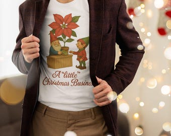 May include: White t-shirt with a Christmas graphic. The design shows a poinsettia plant, a gnome, and an elf, with the text "A little Christmas Business". The shirt is worn with a maroon blazer and brown trousers.