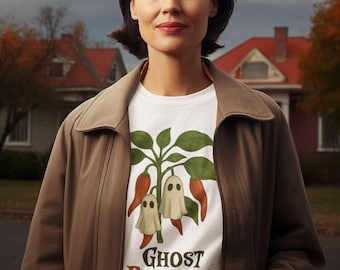 May include: A white t-shirt featuring a graphic illustration of a green pepper plant with orange chili peppers and two white ghost-shaped figures draped over the peppers, with the text "GHOST PEPPERS" printed in a brown serif font below the plant.