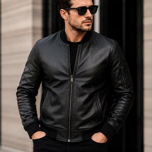 Pode incluir: Um blusão bomber de couro preto com gola, punhos e bainha canelados. O casaco possui um fecho de correr central em metal, dois bolsos laterais e um pequeno bolso utilitário com fecho na manga esquerda, usado sobre uma camisa preta lisa.