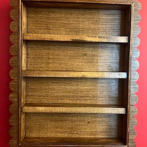 May include: A wooden wall shelf with four horizontal shelves. The frame has a scalloped edge. The wood has a warm brown stain, and the shelves are evenly spaced within the rectangular frame. The shelf is designed for displaying small items.