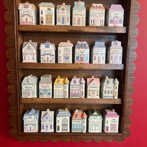May include: A wooden spice rack with a scalloped edge, holding miniature ceramic houses. Each house is labeled with a spice name like "Basil" or "Cinnamon." The houses are painted in pastel colors with detailed architectural features.