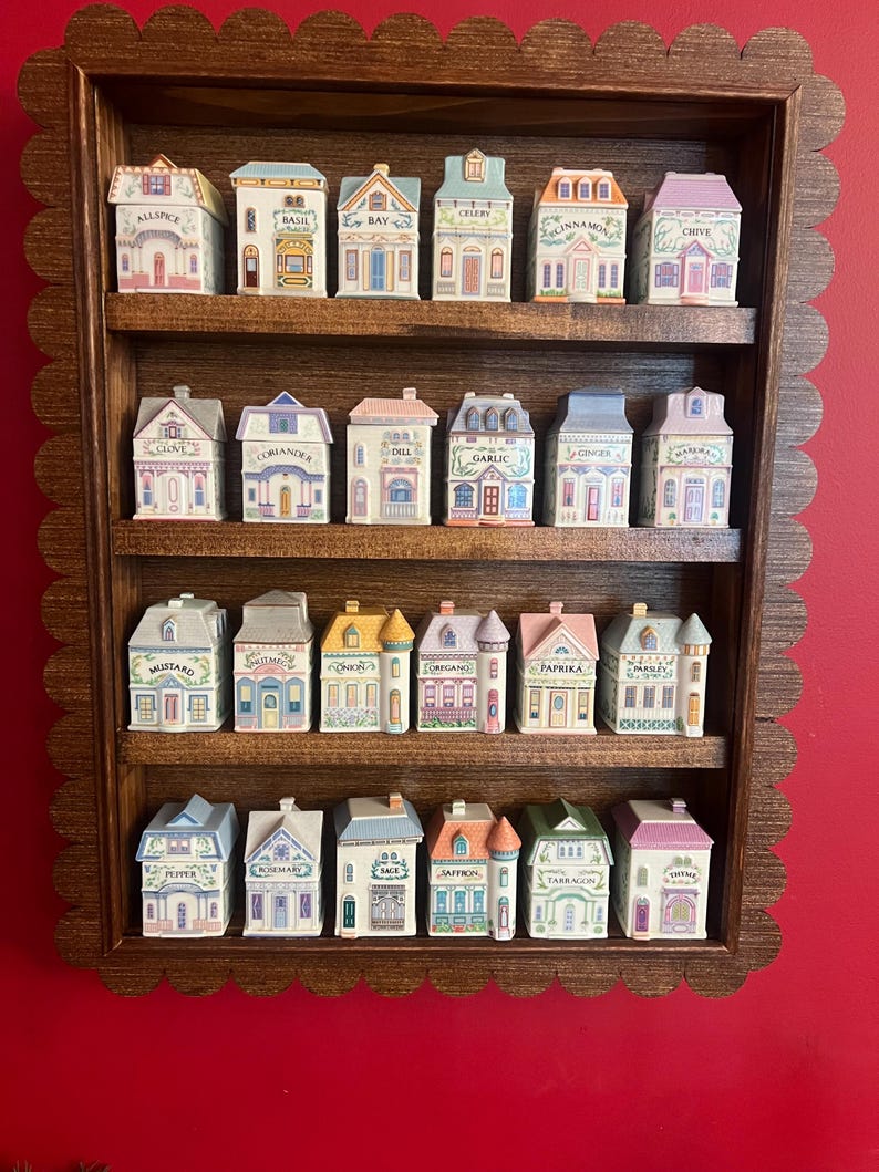 May include: A wooden spice rack with a scalloped edge, holding miniature ceramic houses. Each house is labeled with a spice name like "Basil" or "Cinnamon." The houses are painted in pastel colors with detailed architectural features.