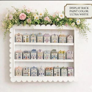 May include: A white display rack with scalloped edges holds miniature, colorful houses. The houses have varying roof colors and architectural details. The rack is adorned with a floral arrangement of pink roses and white daisies. The text on the image reads "DISPLAY RACK PAINT COLOR: ULTRA WHITE."