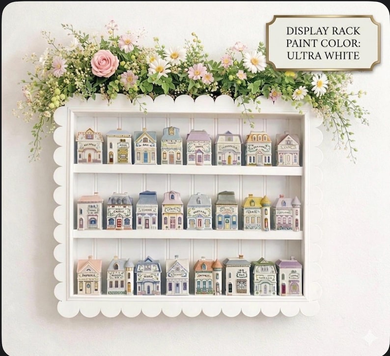 May include: A white display rack with scalloped edges holds miniature, colorful houses. The houses have varying roof colors and architectural details. The rack is adorned with a floral arrangement of pink roses and white daisies. The text on the image reads "DISPLAY RACK PAINT COLOR: ULTRA WHITE."