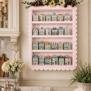 May include: A pink, scalloped-edge shelf displays miniature ceramic houses with colorful details. The houses have varying architectural styles and are arranged on multiple shelves. Floral arrangements adorn the top of the shelf, adding a decorative touch.