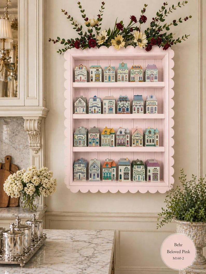 May include: A pink, scalloped-edge shelf displays miniature ceramic houses with colorful details. The houses have varying architectural styles and are arranged on multiple shelves. Floral arrangements adorn the top of the shelf, adding a decorative touch.
