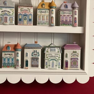 May include: A white, scalloped-edged shelf displays a collection of ceramic spice jars shaped like miniature buildings. Each jar is labeled with a spice name, featuring detailed architectural designs and colorful accents.