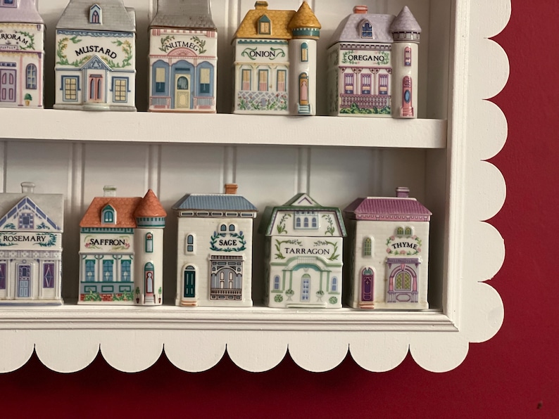 May include: A white, scalloped-edged shelf displays a collection of ceramic spice jars shaped like miniature buildings. Each jar is labeled with a spice name, featuring detailed architectural designs and colorful accents.