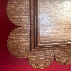 May include: Close-up of a decorative wooden frame with a scalloped edge. The frame has a light brown, textured finish with a darker brown trim. The frame is likely for a mirror or artwork.
