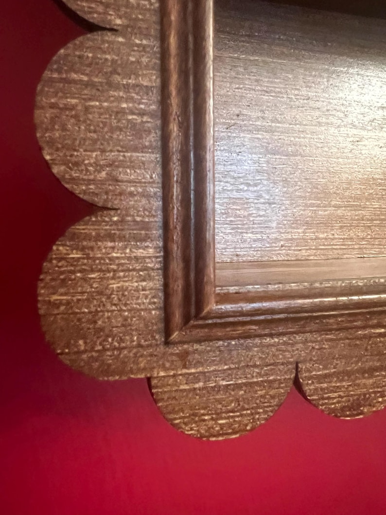 May include: Close-up of a decorative wooden frame with a scalloped edge. The frame has a light brown, textured finish with a darker brown trim. The frame is likely for a mirror or artwork.