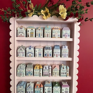 May include: A pink, scalloped-edged shelf displaying miniature ceramic spice jars shaped like colorful houses. The jars are arranged on three shelves, with floral arrangements above. The jars have labels like "Cinnamon" and "Bay".