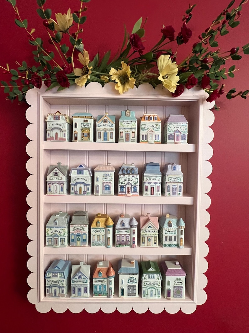 May include: A pink, scalloped-edged shelf displaying miniature ceramic spice jars shaped like colorful houses. The jars are arranged on three shelves, with floral arrangements above. The jars have labels like "Cinnamon" and "Bay".