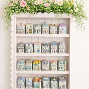 May include: A white scalloped shelf displays miniature ceramic houses with various architectural styles and pastel colors. The shelf is adorned with a floral arrangement of pink and yellow flowers and greenery, creating a charming, decorative display.