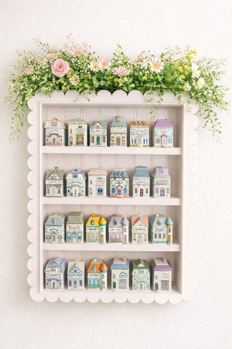 May include: A white scalloped shelf displays miniature ceramic houses with various architectural styles and pastel colors. The shelf is adorned with a floral arrangement of pink and yellow flowers and greenery, creating a charming, decorative display.