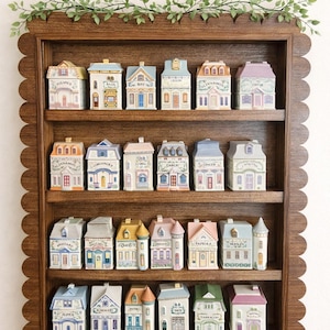 May include: A wooden shelf with a scalloped edge displays miniature ceramic houses, each with a unique design and labeled with spice names like "Bay" and "Cinnamon." A green vine decorates the top of the shelf. The houses are in various colors.