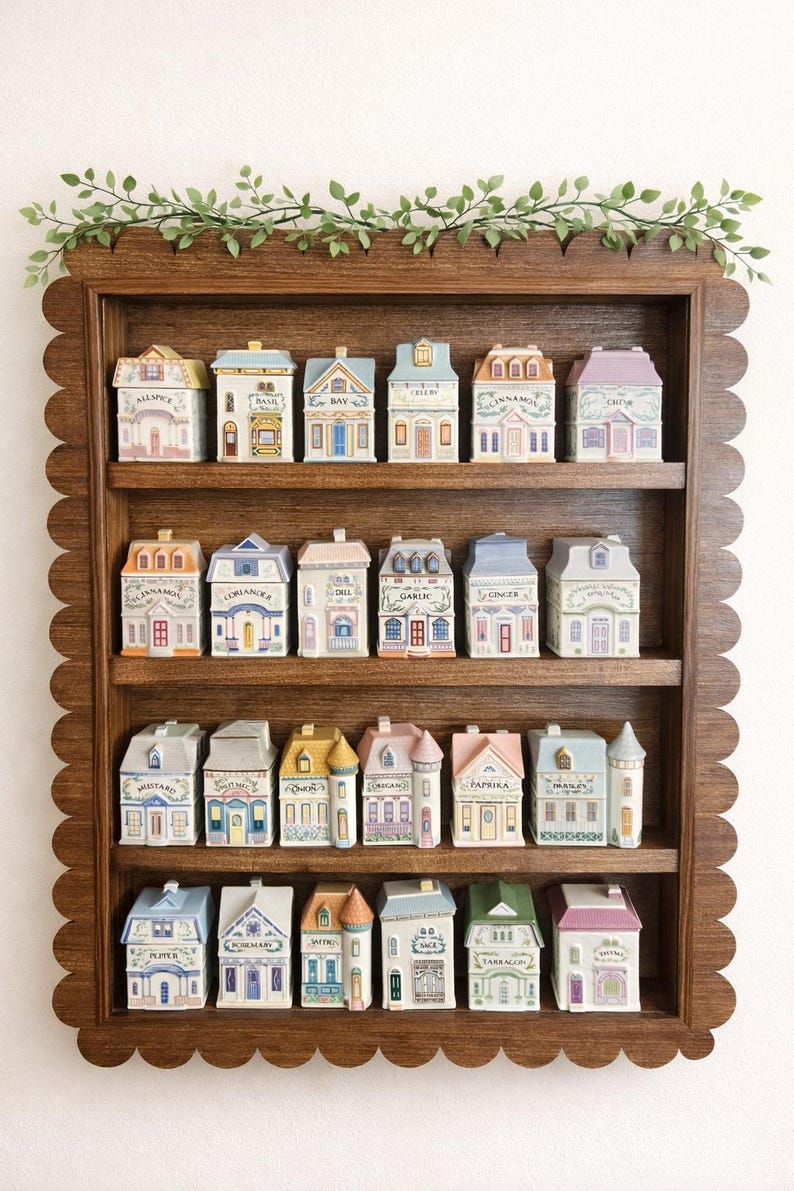 May include: A wooden shelf with a scalloped edge displays miniature ceramic houses, each with a unique design and labeled with spice names like "Bay" and "Cinnamon." A green vine decorates the top of the shelf. The houses are in various colors.