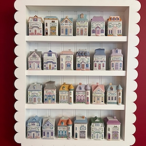 May include: A white, scalloped-edged shelf unit displays a collection of miniature ceramic spice jars shaped like houses. Each jar is labeled with a spice name in black text, and the houses are painted in various colors with detailed architectural features.