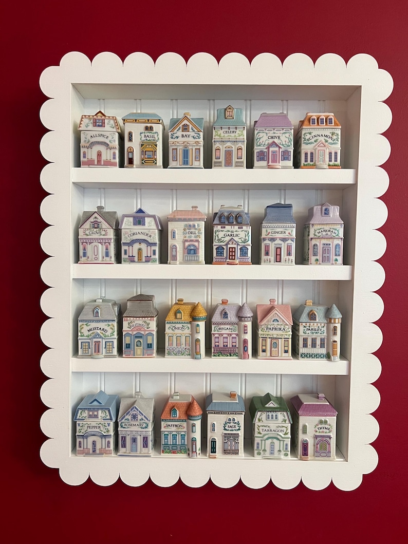 May include: A white, scalloped-edged shelf unit displays a collection of miniature ceramic spice jars shaped like houses. Each jar is labeled with a spice name in black text, and the houses are painted in various colors with detailed architectural features.