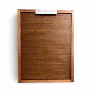 May include: A rectangular wooden clipboard with a dark brown surface and a lighter brown frame. A silver metal clip is attached to the top. The clipboard is designed for holding papers and documents.