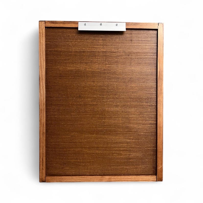 May include: A rectangular wooden clipboard with a dark brown surface and a lighter brown frame. A silver metal clip is attached to the top. The clipboard is designed for holding papers and documents.