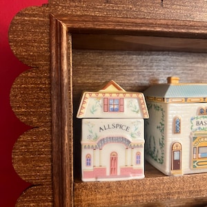 May include: A wooden spice rack with a scalloped edge, holding several ceramic spice jars. The front jar is labeled "ALLSPICE" and is shaped like a house with pink and white details. Other jars are visible.