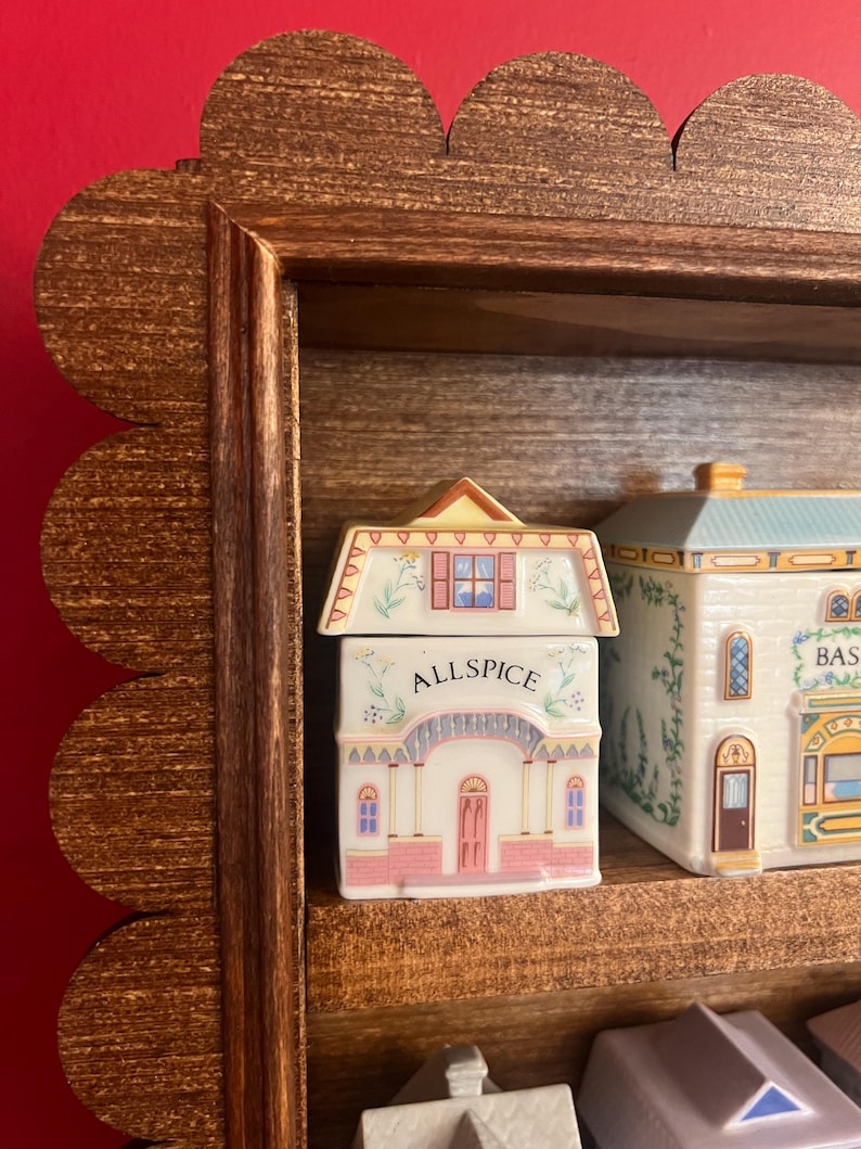 May include: A wooden spice rack with a scalloped edge, holding several ceramic spice jars. The front jar is labeled "ALLSPICE" and is shaped like a house with pink and white details. Other jars are visible.