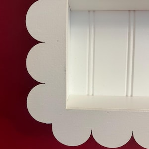 May include: A white, scalloped-edge shelf with a recessed interior. The shelf has a rectangular opening with vertical paneling. The frame is painted white, and the background is a solid red.