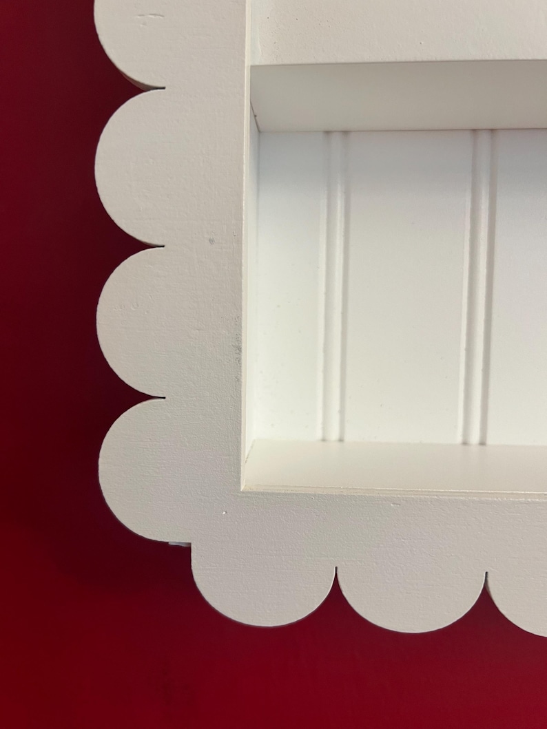 May include: A white, scalloped-edge shelf with a recessed interior. The shelf has a rectangular opening with vertical paneling. The frame is painted white, and the background is a solid red.