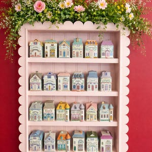 May include: A pink scalloped shelf displays miniature ceramic houses with various architectural styles and colors. Each house has a unique design and label. The shelf is adorned with a floral arrangement of pink and white flowers and greenery against a red background.