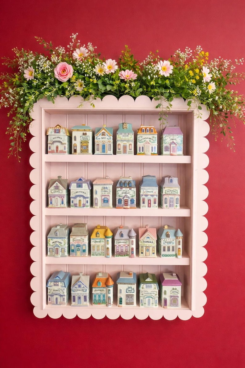 May include: A pink scalloped shelf displays miniature ceramic houses with various architectural styles and colors. Each house has a unique design and label. The shelf is adorned with a floral arrangement of pink and white flowers and greenery against a red background.