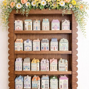May include: A wooden shelf with a scalloped edge displays miniature ceramic houses, each with unique architectural details and labels. The shelf is adorned with a floral arrangement of various flowers. The houses are arranged in rows, creating a charming display.