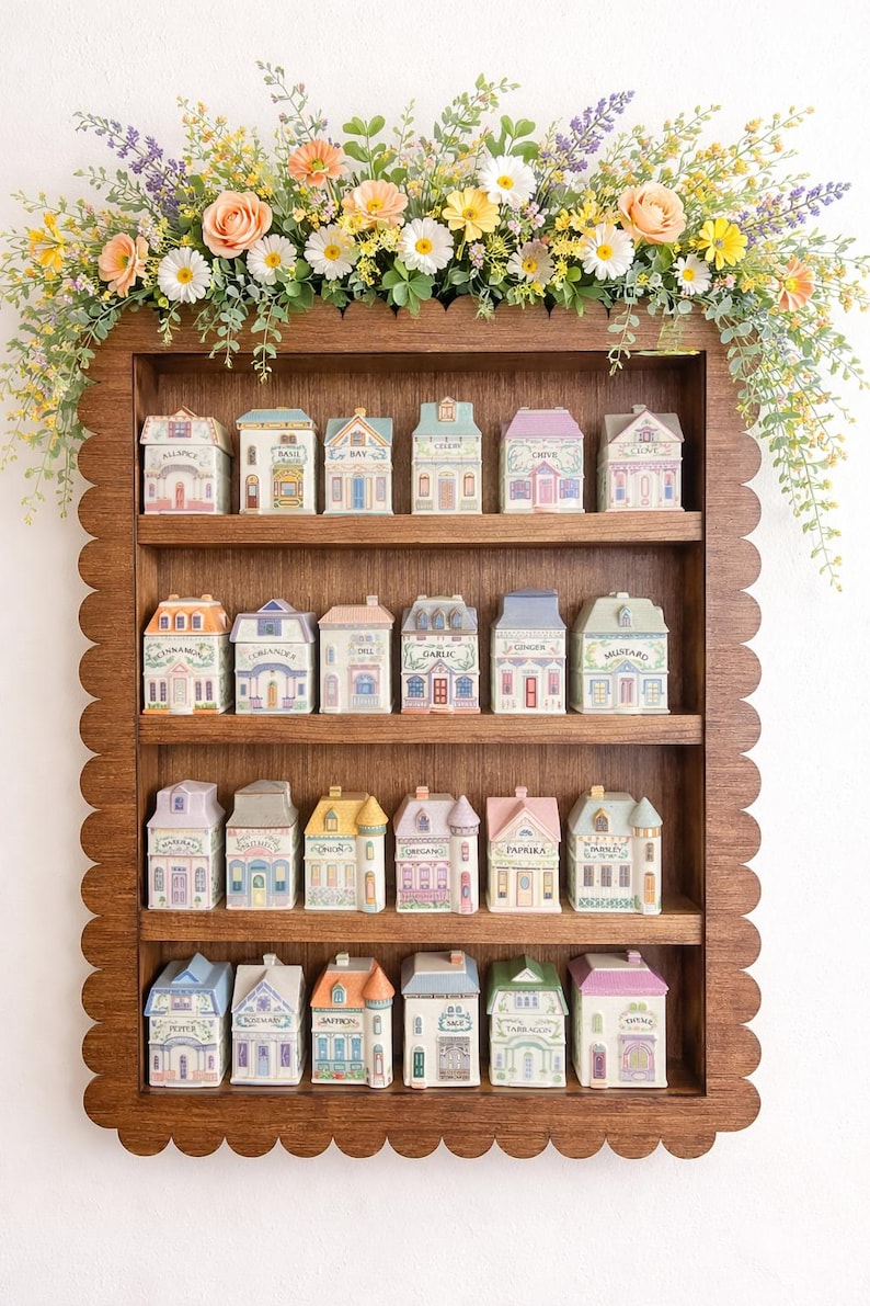 May include: A wooden shelf with a scalloped edge displays miniature ceramic houses, each with unique architectural details and labels. The shelf is adorned with a floral arrangement of various flowers. The houses are arranged in rows, creating a charming display.