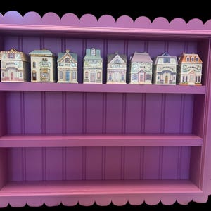 May include: A decorative, scalloped-edge, purple spice rack with three shelves. The top shelf holds eight miniature house-shaped spice jars, each labeled with a different herb or spice name. The rack is painted a vibrant purple.