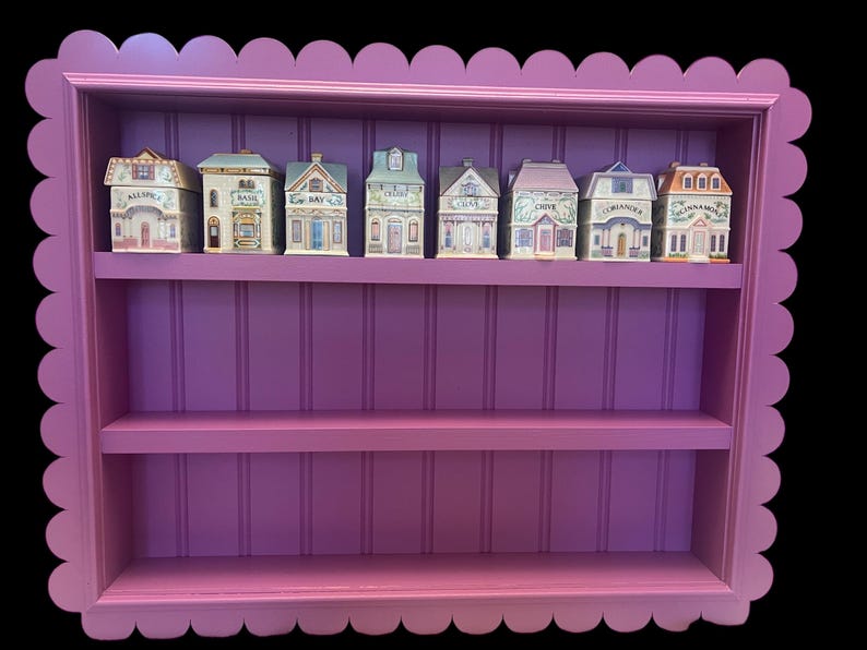 May include: A decorative, scalloped-edge, purple spice rack with three shelves. The top shelf holds eight miniature house-shaped spice jars, each labeled with a different herb or spice name. The rack is painted a vibrant purple.