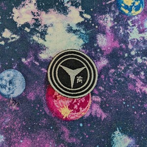 May include: A circular embroidered patch featuring a black background with concentric white rings and a central white three-pointed star emblem. The patch is placed on a fabric surface with a colorful galaxy print of purple, blue, and pink nebulae.